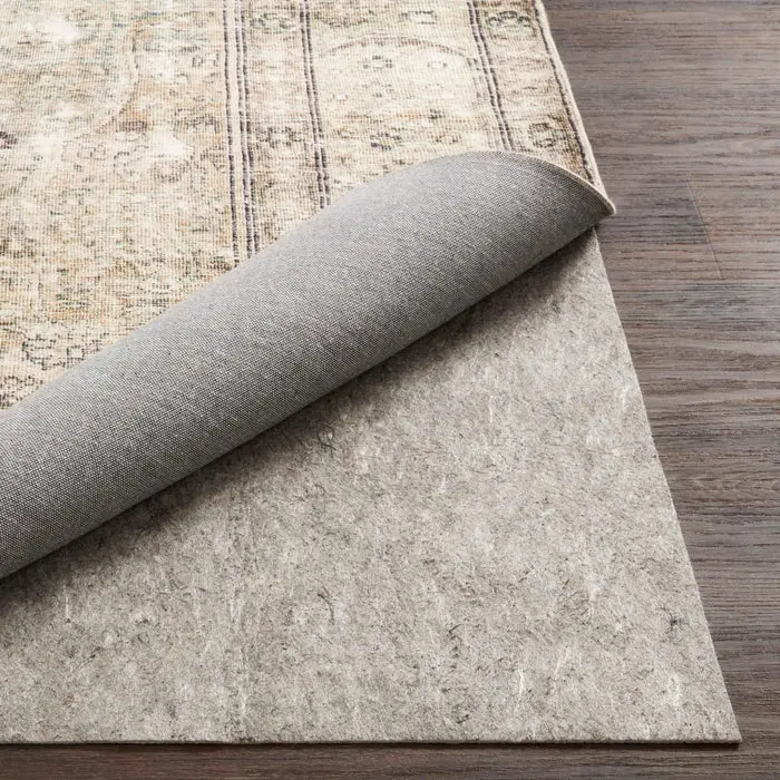 Corner of a patterned area rug peeled back to show a grey rug pad underneath on dark wood flooring.